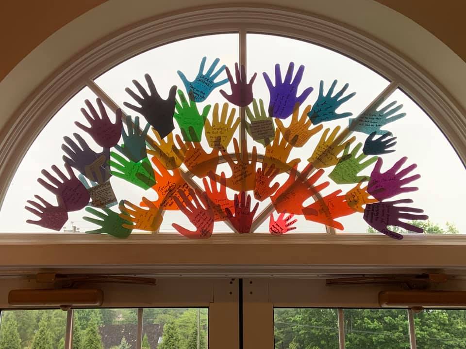Colorful hand-shaped papers with messages are displayed on a semi-circular window, overlooking greenery outside. The scene is artistic and uplifting.
