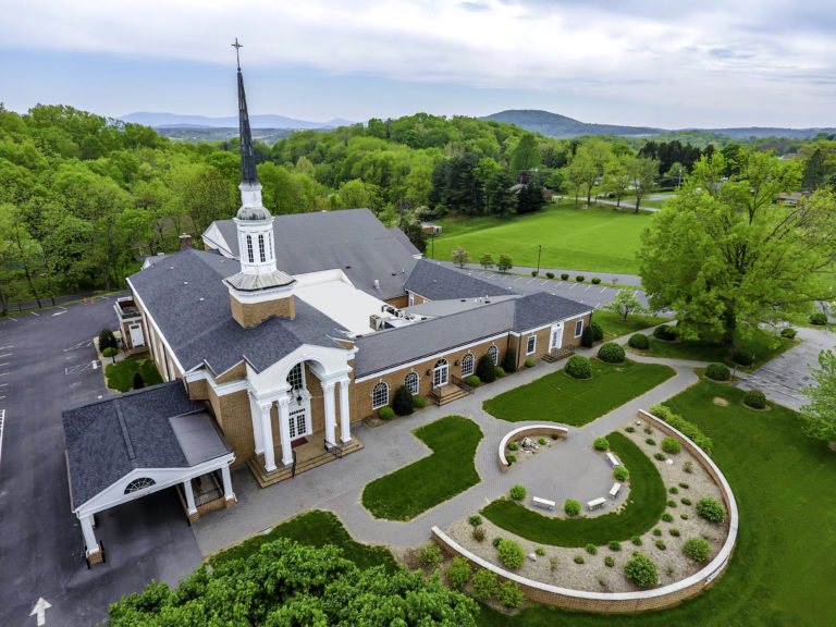 Aerial view of a large brick church with a prominent steeple, surrounded by green lawns and trees, against distant rolling hills.