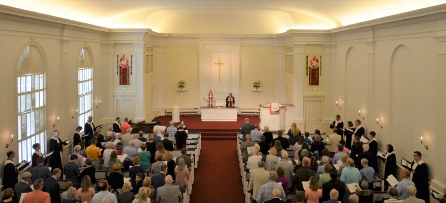 A congregation gathers inside a brightly lit church with decorated walls, a central altar, and high ceilings, observing a service in progress.