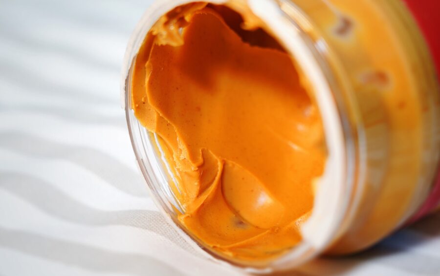 An open jar of smooth orange-colored spread, possibly cheese or nut butter, with visible creamy texture inside, resting on a white surface.