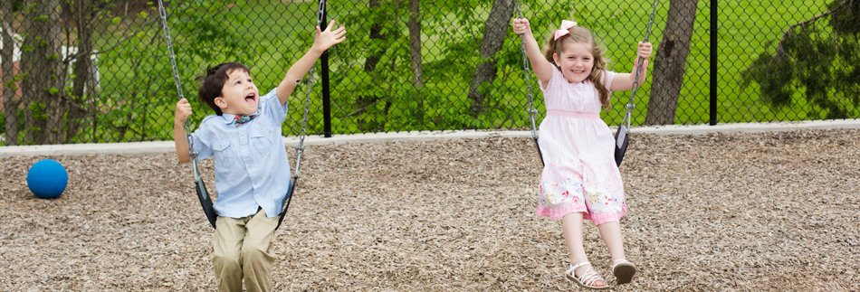 Two children joyfully swinging at a playground. A blue ball rests on the ground. Greenery and a chain-link fence are in the background.
