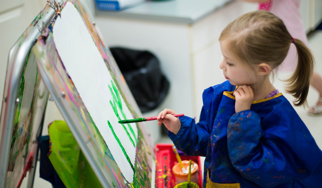 A child in a blue smock paints on an easel with focused expression, surrounded by art supplies in a bright room.