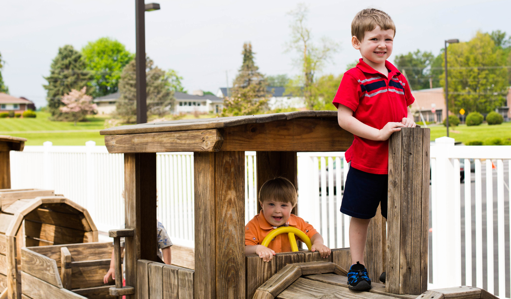 Children play joyfully on a wooden playground train surrounded by greenery and a white fence on a bright day.
