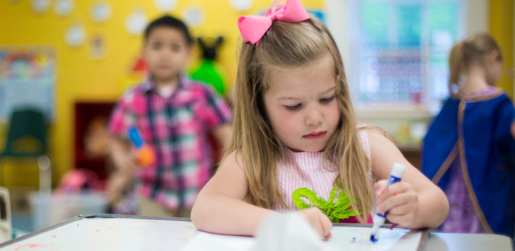 A young child focuses on drawing with a marker in a colorful classroom. Two other children play in the background.
