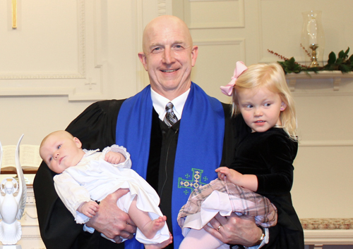 A person dressed in a robe holds a baby and a toddler inside a church setting, featuring religious decorations and furnishings.