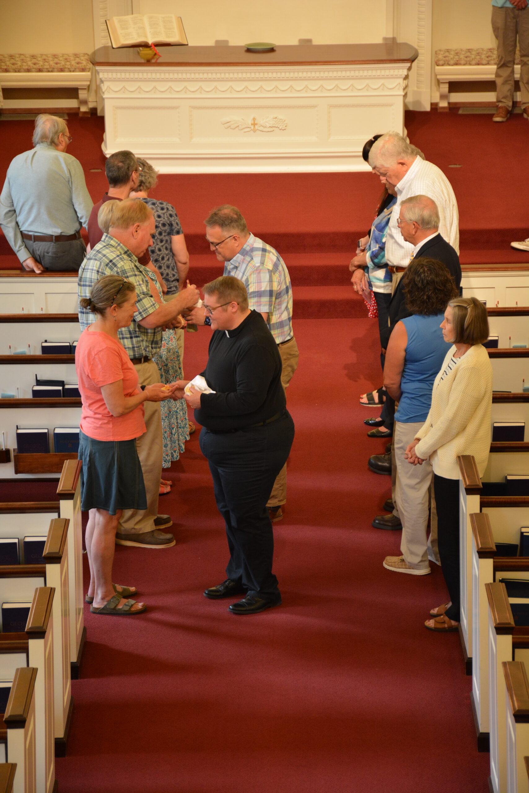 People in a church engage in a line ceremony, with a person in black attire interacting with others. Altar visible in background.