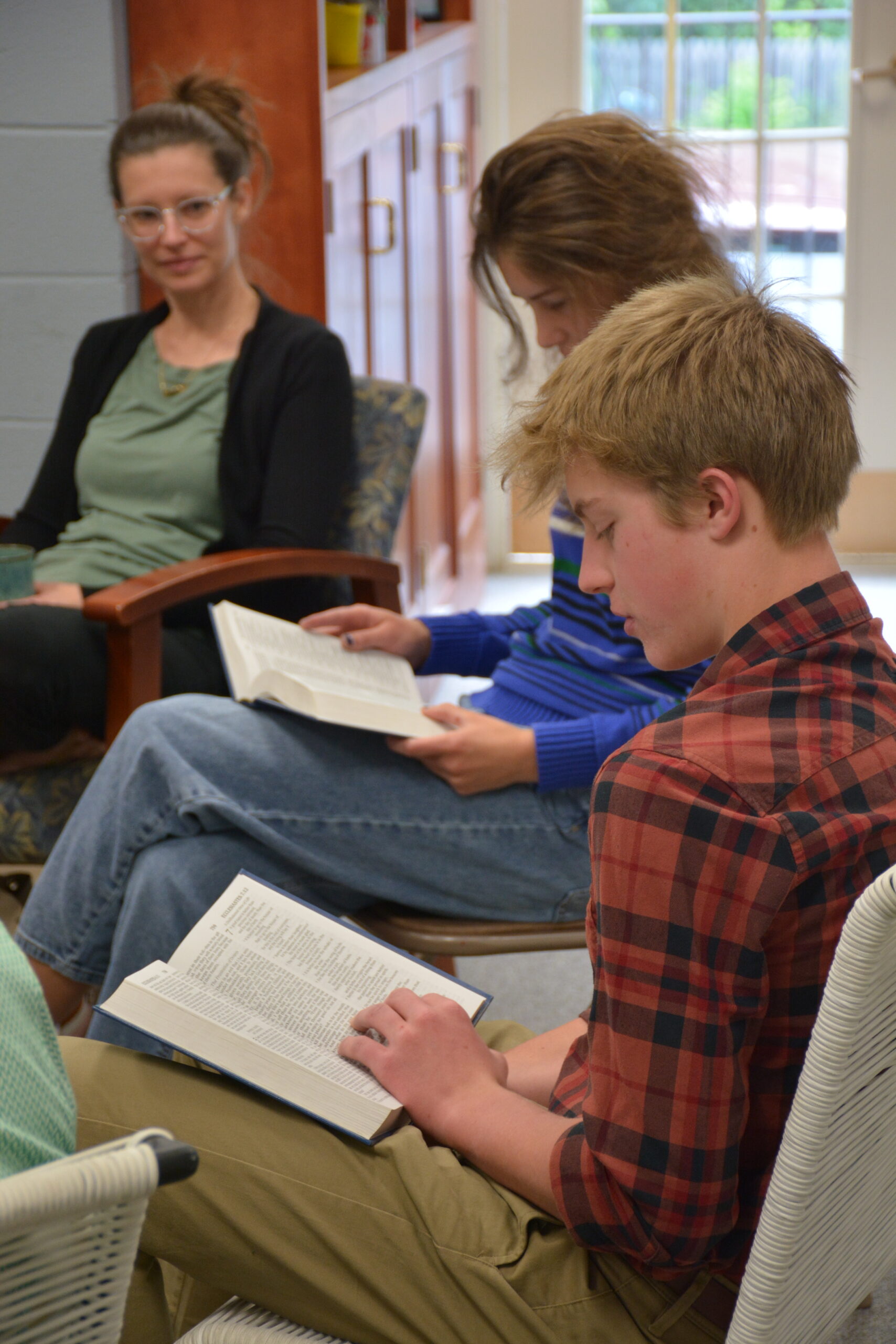 Three people are seated, reading books in a cozy room lit by natural light. The focus is on learning and concentration.
