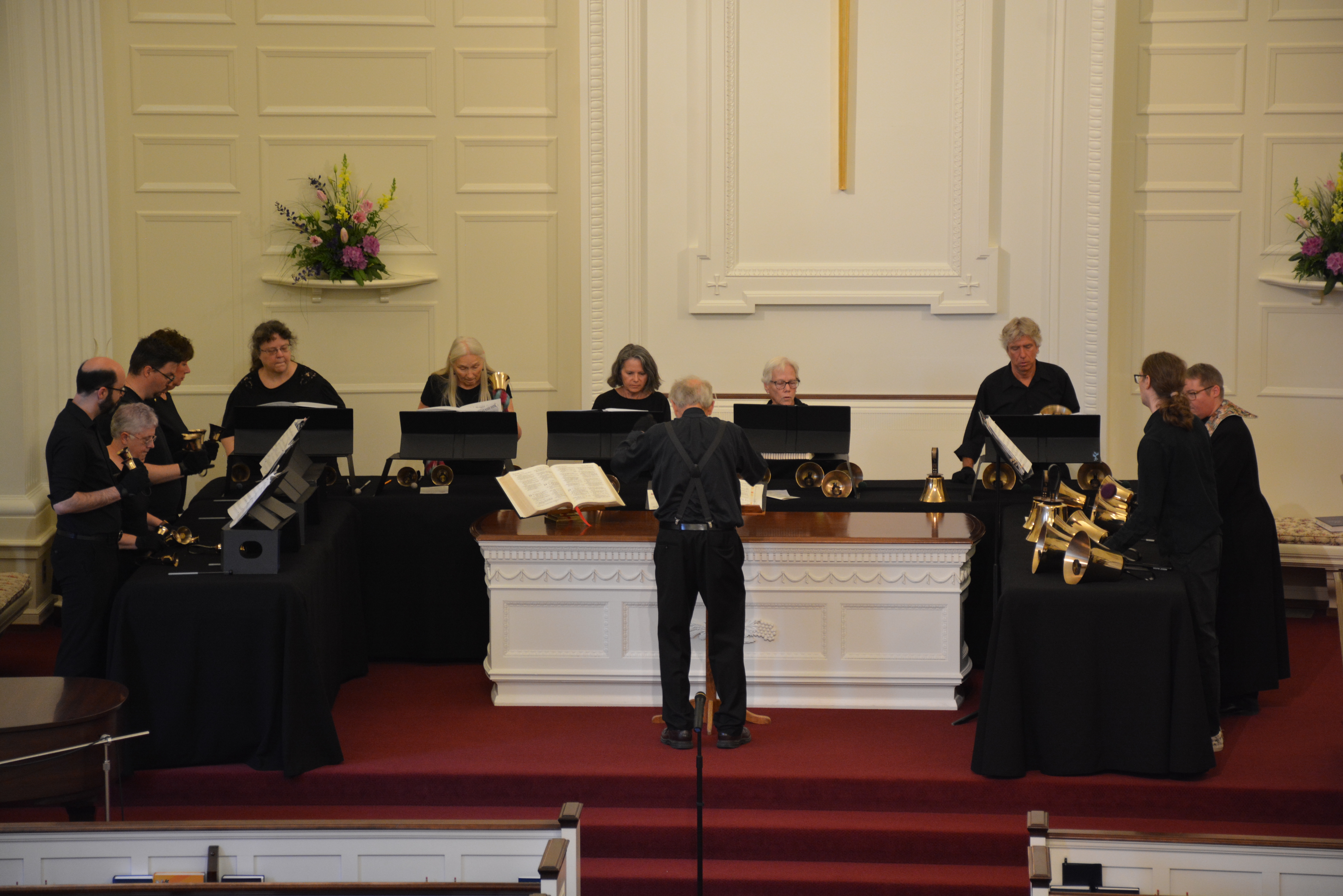 A group of people, dressed in black, perform with handbells on a stage inside a church with a prominent cross and flowers.