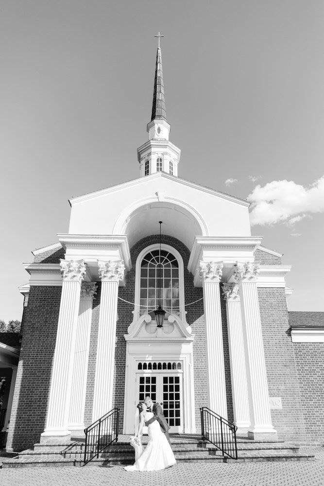A couple poses affectionately outside a large, columned church, with a tall spire above. The scene is captured in black and white.