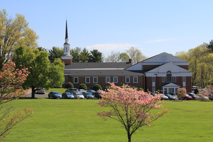 A large brick building with a steeple stands amidst green lawns and blooming trees, with cars parked nearby under a clear sky.