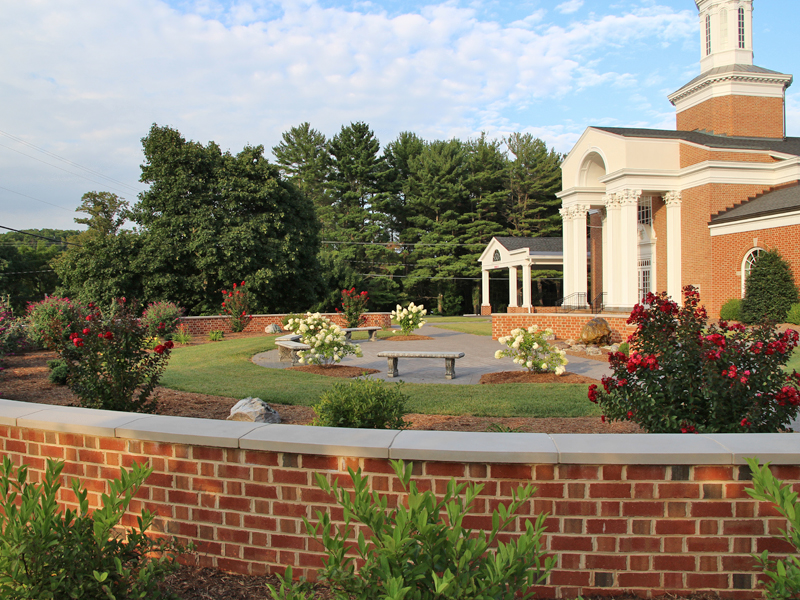 A brick building with columns, surrounded by landscaped gardens and benches, features in a serene setting under a partly cloudy sky.