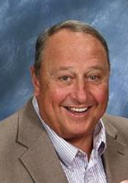 A person with a warm smile, wearing a suit jacket and shirt, is photographed against a blue, textured background.