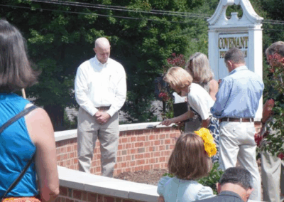 Several people gather around a brick structure near a Covenant Presbyterian Church sign, appearing to participate in a solemn outdoor ceremony or event.