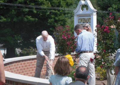 A person shovels dirt at a community event beside a sign reading "Covenant." Several people observe the activity near a brick structure and foliage.
