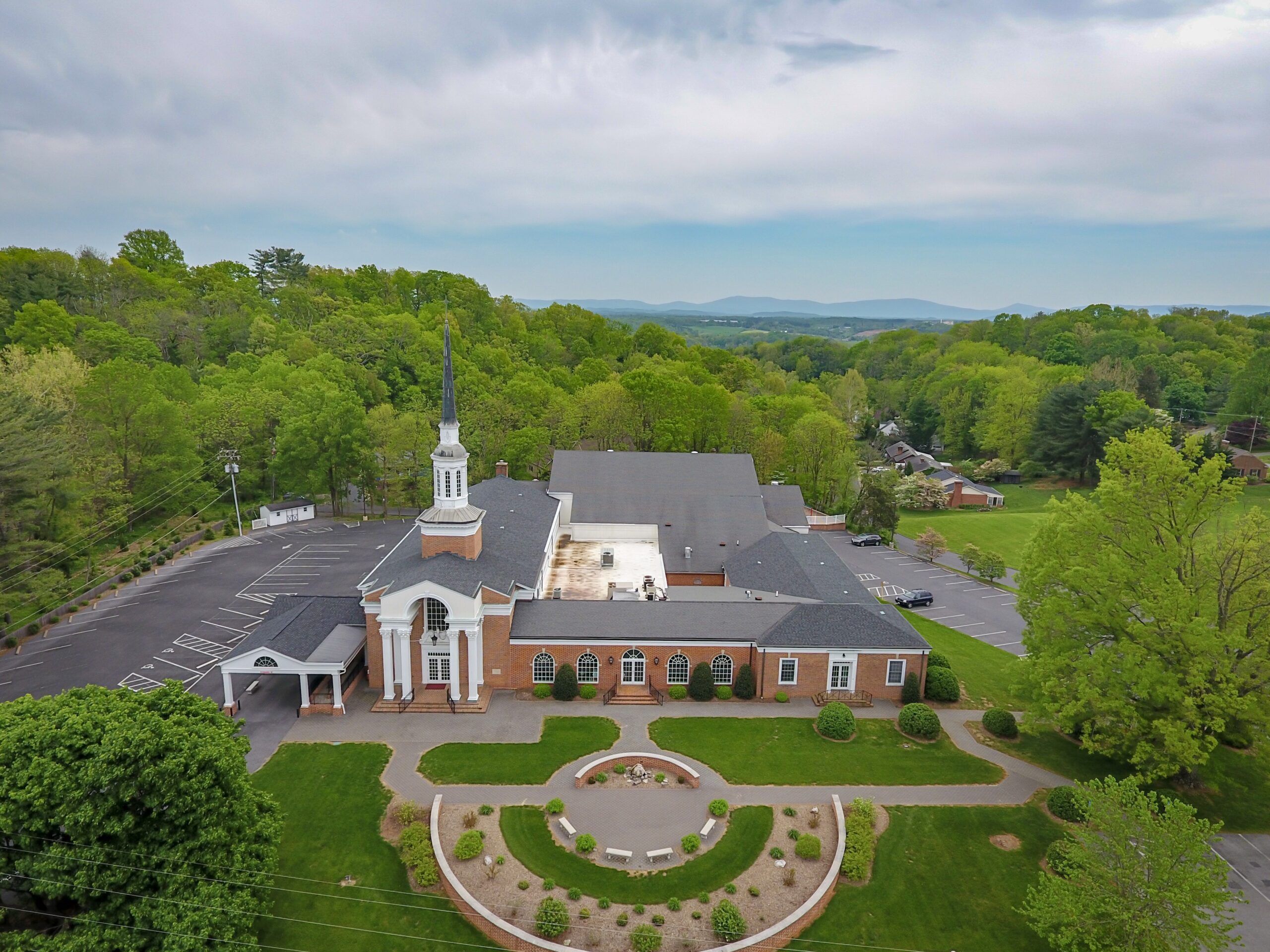 Aerial view of a church with a tall steeple, surrounded by lush green trees and scenic hills in the background.