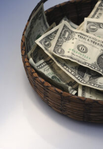 A woven basket filled with assorted U.S. dollar bills sits on a smooth, light-colored surface, symbolizing monetary collection or donation.